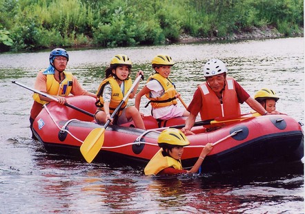 ヘルメットを被りライフジャケットを着てゴムボートに乗り川下りを体験している子ども4人と2人の男性の写真