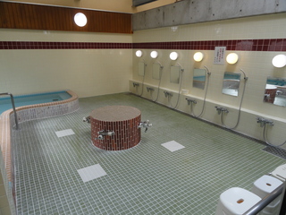 右手前に重なっている椅子と右奥の壁に沿って並んだ鏡が付いている6つのシャワー台があり、左側に浴場がある浴場の写真