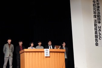 発表者と書かれた檀上に6名の学生が横一列に並んで立ち報告を行っている成果報告会の様子の写真