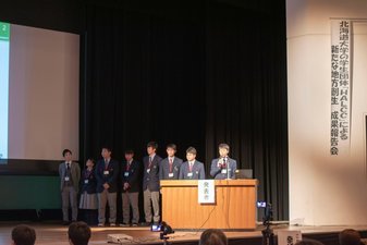 ステージのスクリーンに資料が映しだされた右側の発表者と書かれた檀上に男性教師と7名の高校生が横一列に並んで立ち一番右側に立っている男子学生が報告を行っている様子の写真