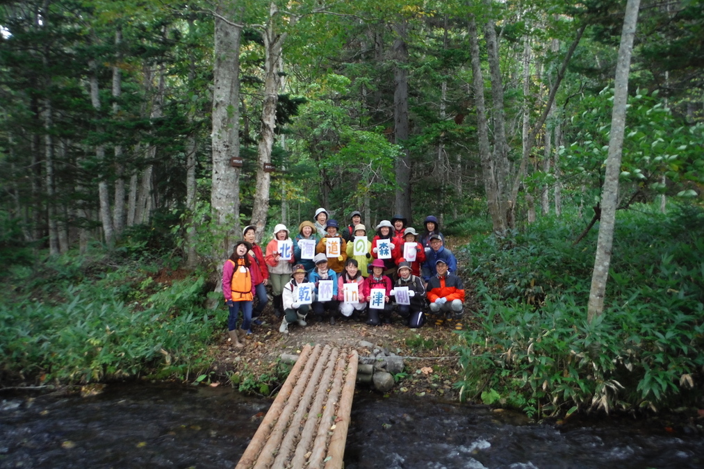 森林の中を走る小川を前に、記念撮影に応じる参加者たちの集合写真。参加者の手には「北海道の自然に乾杯イン津別」と書かれたメッセージパネルが確認できる