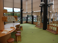 緑色のカーペットの敷かれた床の上にいくつかの椅子や机があり木でできた収納棚が置いているつべつ木材工芸館キノスの内装の写真