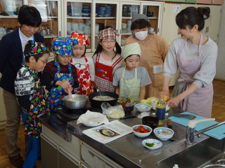 エプロンと三角巾を子供たちと大人たちが調理の様子を見ている写真