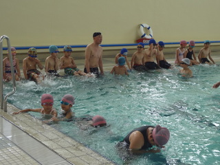 水着姿で帽子を被っている水泳教室の生徒達がプールサイドに腰かけており、泳いでいる生徒と指導をしている講師の先生がプールに入っている水泳教室の様子の写真
