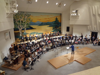 指揮者を前に弓型に配置された演奏中の楽団の写真