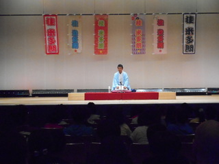 名前が書かれた布が下げられて舞台上で、和服姿で正座し落語をしている男性の写真