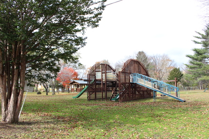 左手前や奥の広葉樹や針葉樹に囲まれた中央にすべり台や階段などが組み合わせられた遊具のある公園の写真